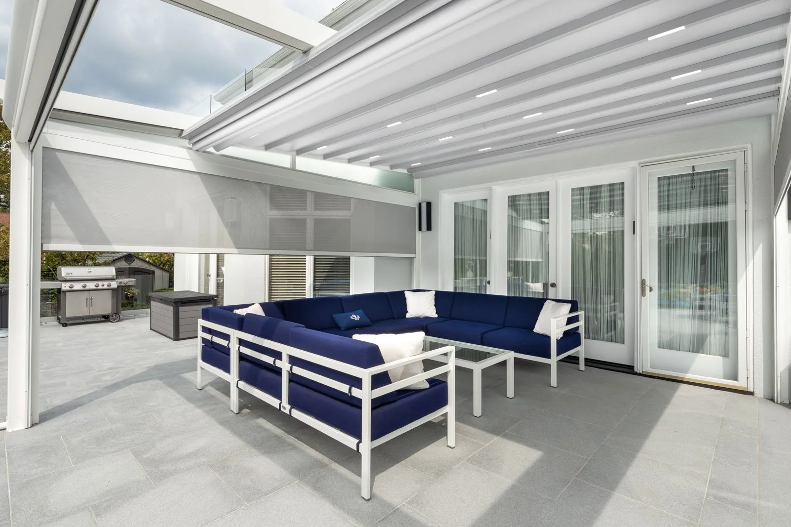 White-framed retractable fabric roof closed over a poolside lounge, navy seating beneath, pool and loungers beyond the open side — photograph