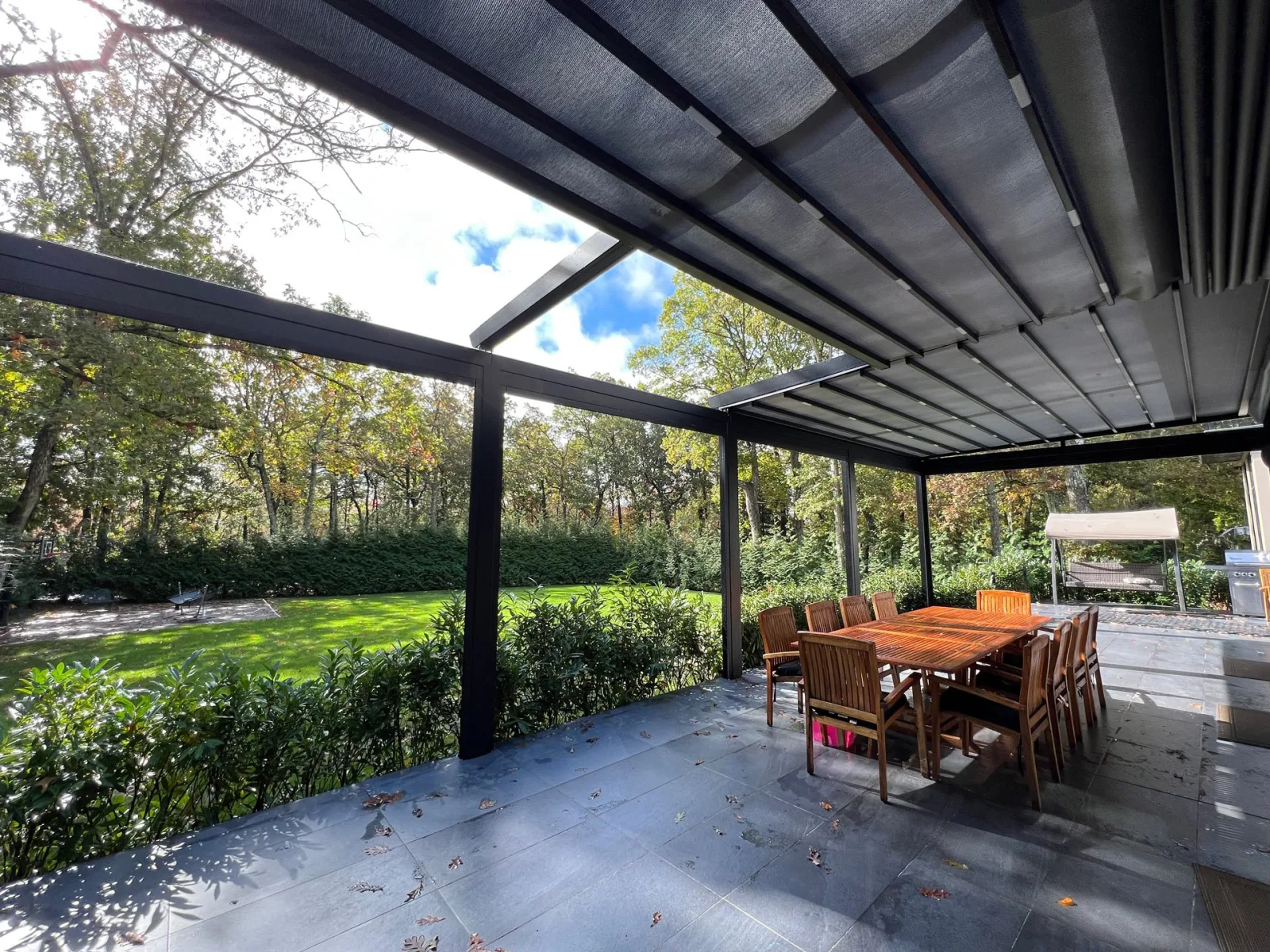 Dark-framed retractable fabric roof caught mid-run over a bluestone patio, open sky ahead of the front bar and taut fabric behind, teak dining table and autumn woods beyond — photograph