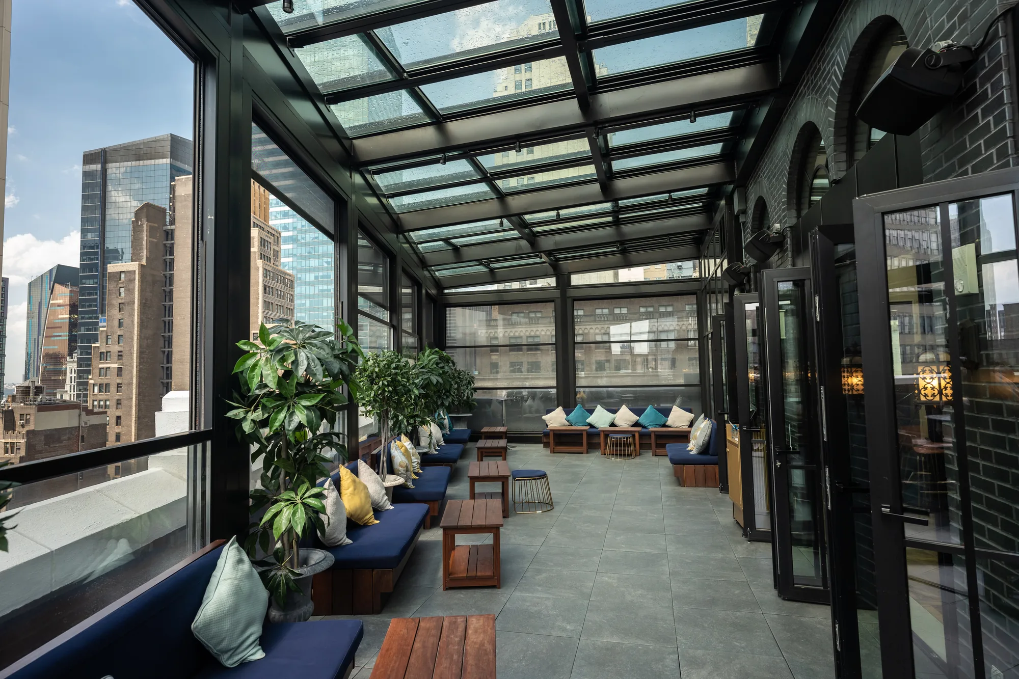 Black-framed glass roof running the length of a rooftop terrace, glazed on the long side with city towers beyond and banquette seating below — photograph