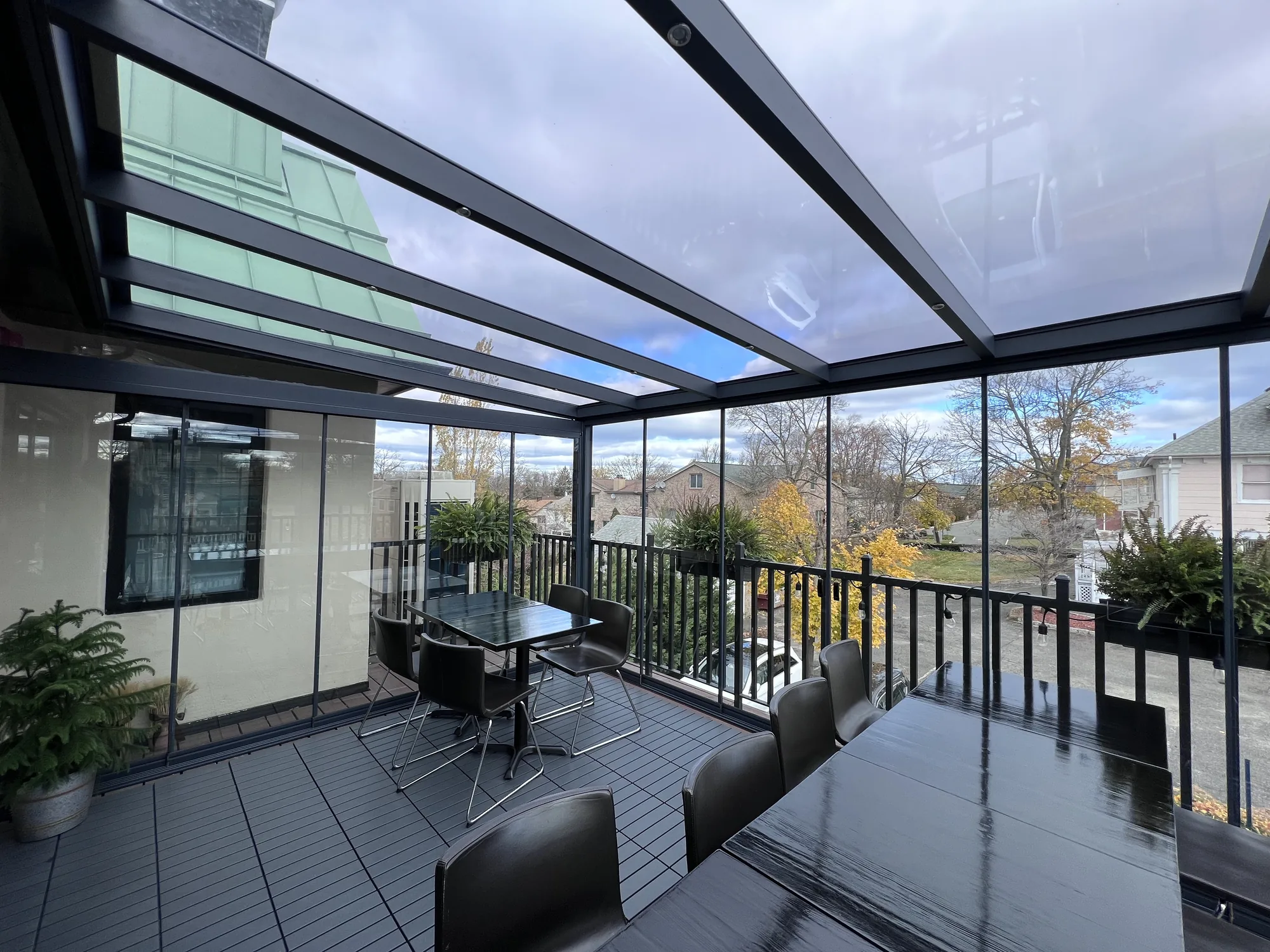 Glass-roofed room on a raised deck with glass side walls, dark tables and chairs inside and an autumn suburban view beyond — photograph