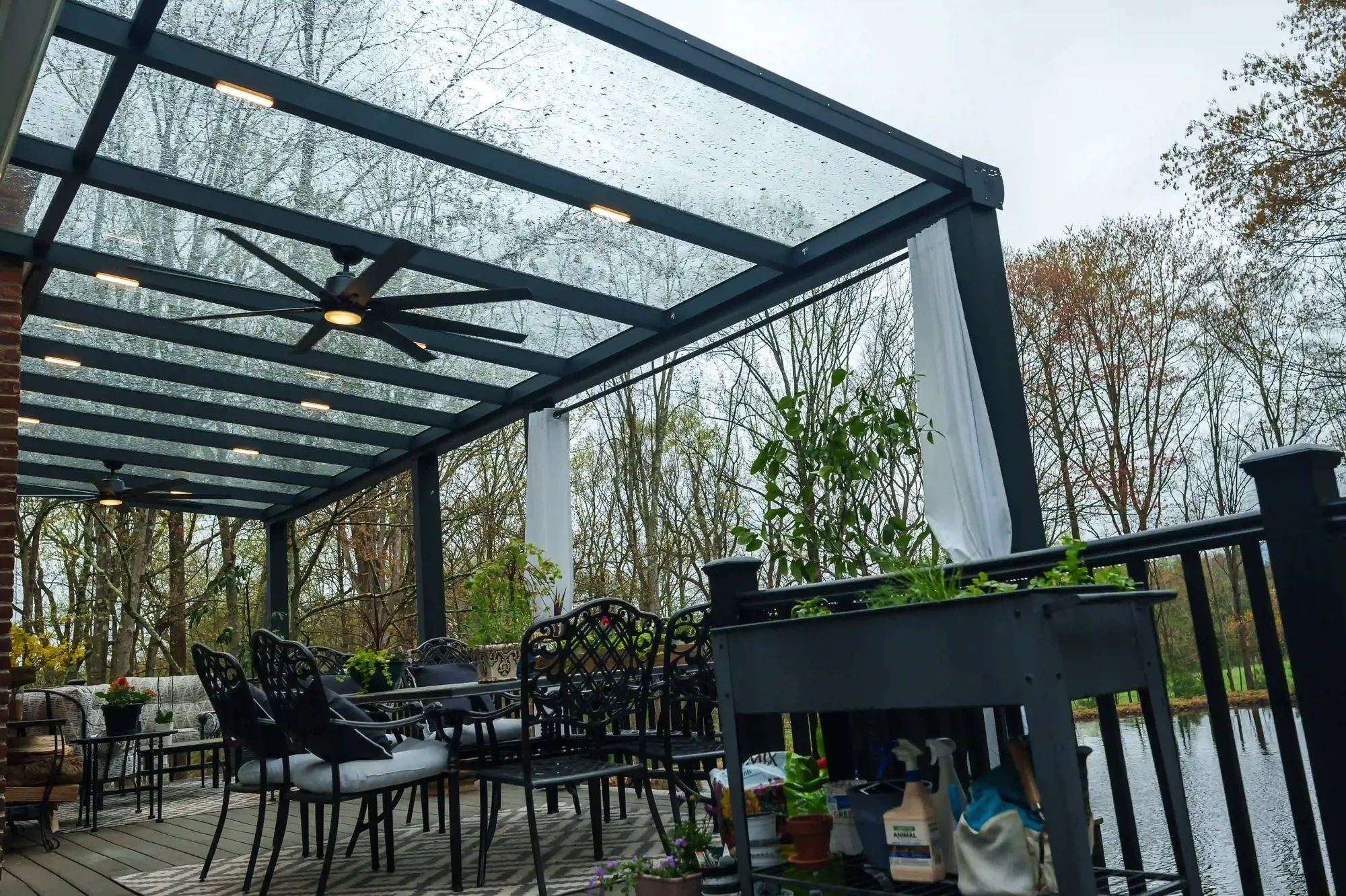 Rain beaded across a glass pergola roof over a waterside deck, black ceiling fans and white curtains beneath, bare trees and water beyond — photograph