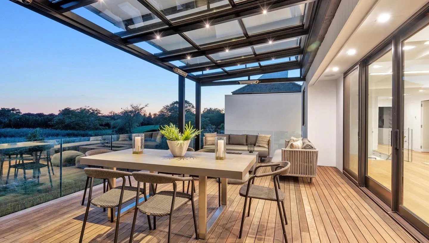 Under a black-framed glass roof on an upper terrace at dusk, LEDs lit in the rafters and a dimming sky through the panes — photograph