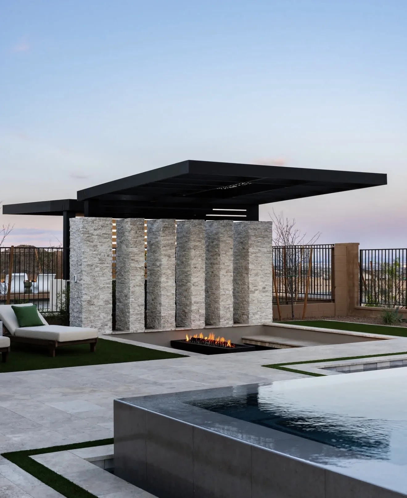 Hard top roof plane carried on a stacked stone wall with lit vertical slots, fire trough and pool below at dusk — photograph