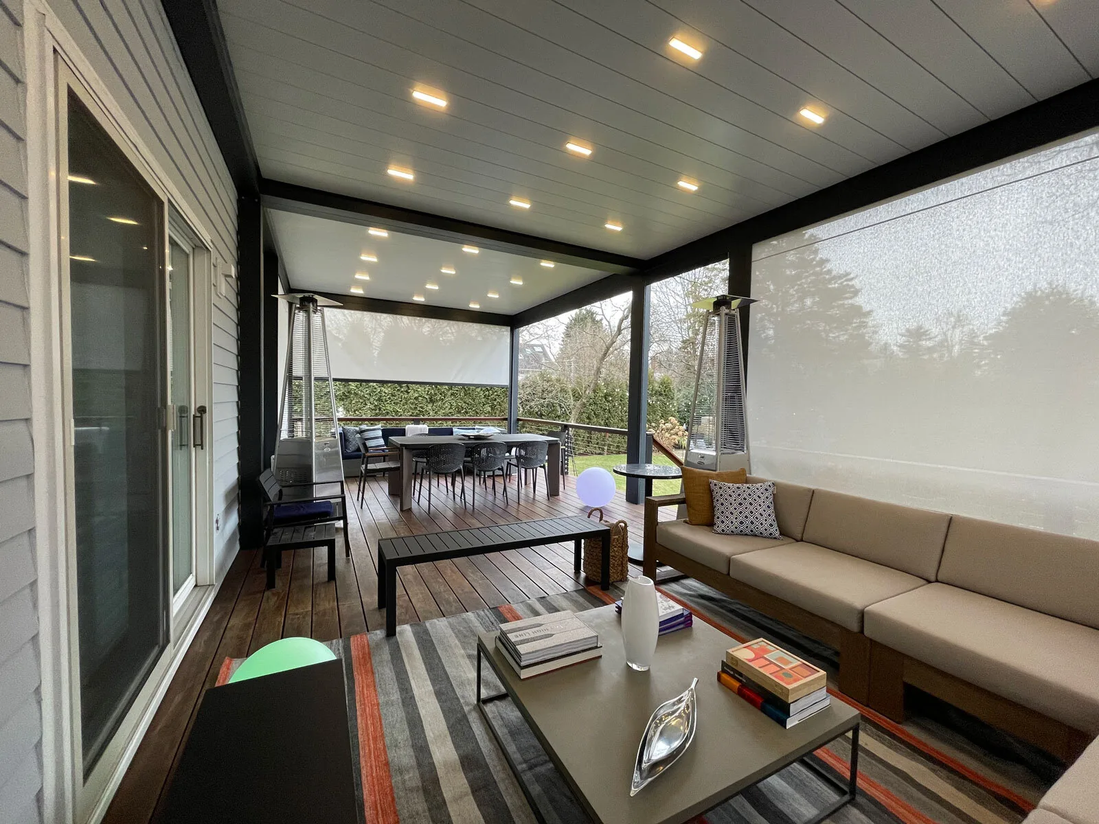 Inside a black-framed louvered roof on a wood deck, blades closed with downlights lit and patio heaters at the posts — photograph