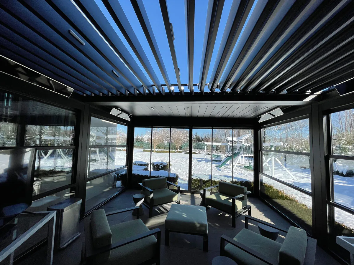 Inside a glazed room under black louvered blades turned open to a blue winter sky, infrared heaters on the beams and snow beyond the glass — photograph