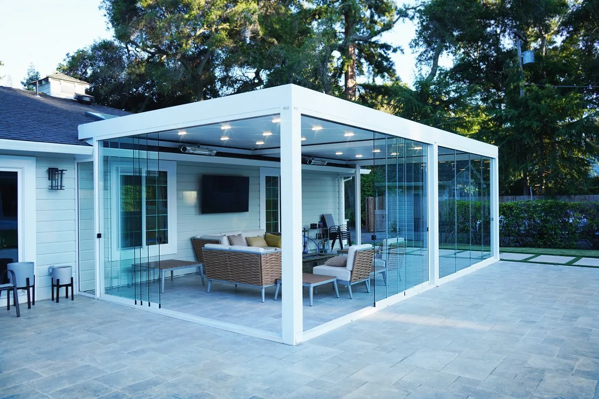 A white-framed freestanding pergola glazed on all sides with sliding panels closed, seating visible inside, on a paved terrace — photograph