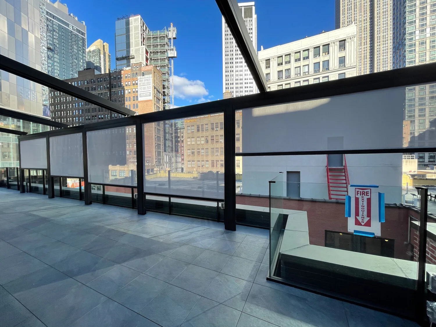 A dark-framed rooftop structure between city towers with motorized screens lowered in their side channels, glass balustrade beyond — photograph