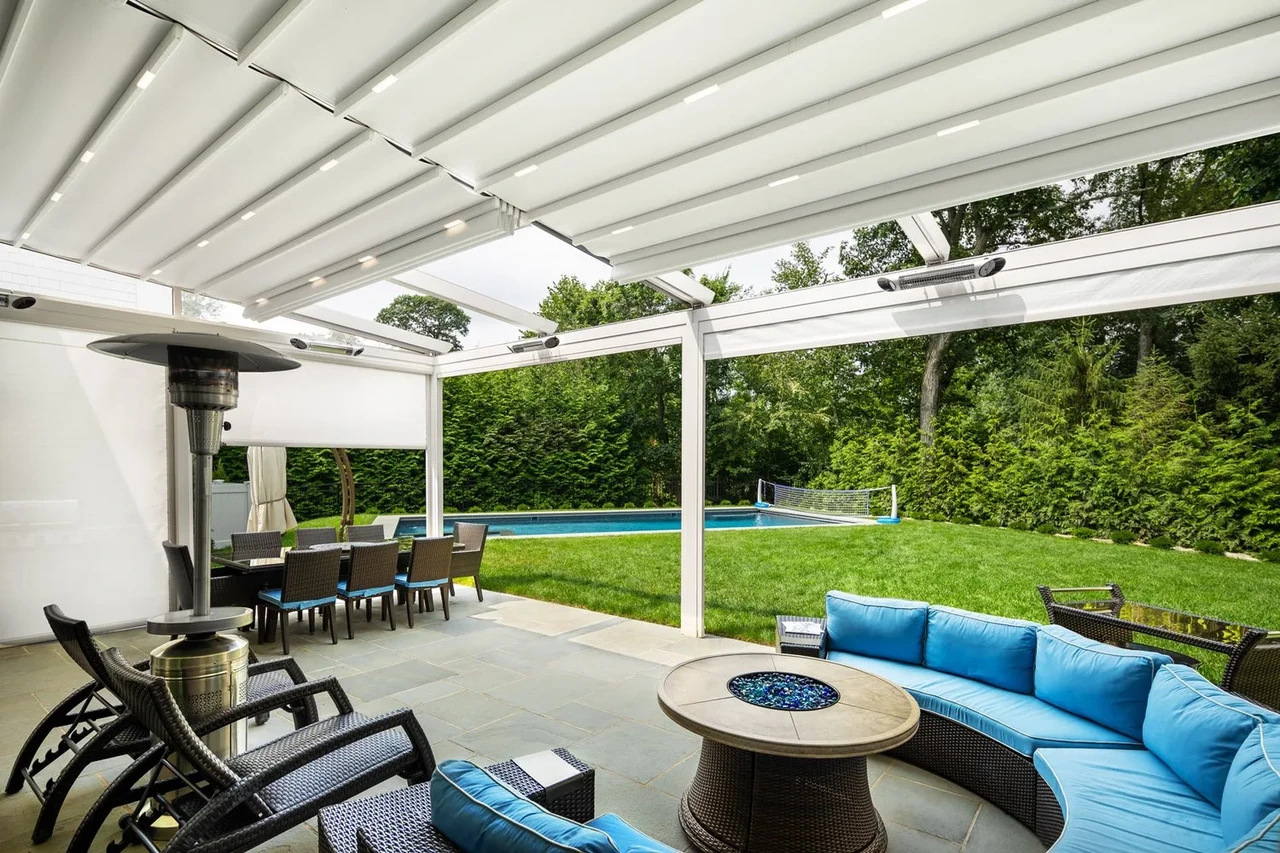 A wide view beneath a white fabric roof from a fire table toward a pool and lawn.