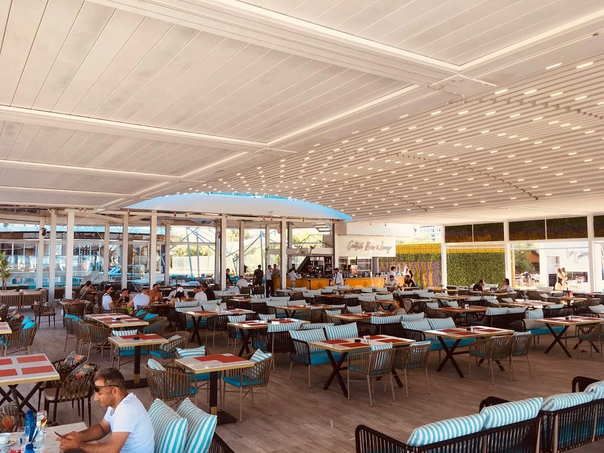 A bright restaurant terrace beneath white fabric roof sections with closely spaced support profiles.