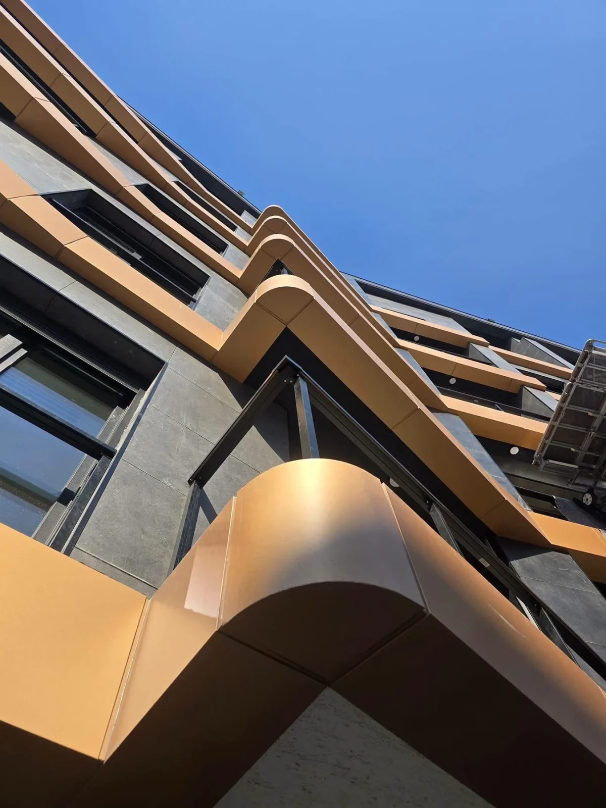 Upward view of curved bronze-colored facade bands and dark stone surfaces beside construction scaffolding.